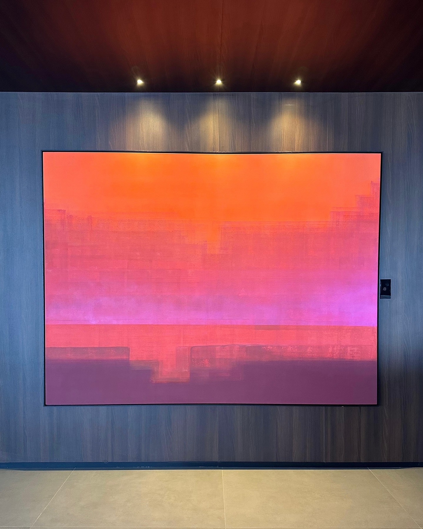 Pintura abstrata em degradê de laranja, rosa e roxo, exposta em parede de madeira escura sob spots de luz